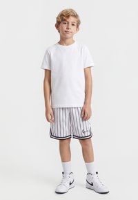 Blond chłopiec stojący, ubrany w gładką białą koszulkę, białe szorty z czarnymi pionowymi paskami, białe skarpety sportowe i białe buty Nike.