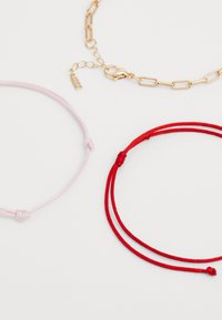 Three bracelets: one pink string with a knot, one red string with two strands, and one gold chain with a lobster clasp and tag.