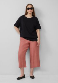 T-shirt en coton noir, coupe décontractée, associé à un pantalon culotte en crêpe corail et des chaussures noires à enfiler avec des accents cloutés. Modèle portant des lunettes de soleil noires.