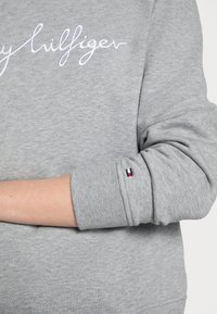 Grijze sweatshirt met ribgebreide manchetten, voorzien van witte borduursel "Tommy Hilfiger". De mouw heeft een kleine vlagaccent in rood, wit en blauw.