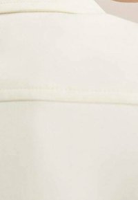 Crème-kleurige stof met een gladde textuur en een subtiele glans; beschikt over een naadaccent bij de halslijn, wat zorgt voor een minimalistisch ontwerp.