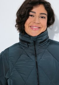 Giacca imbottita in verde teal scuro con colletto alto e zip frontale. Caratterizzata da un motivo a rombi e una superficie liscia e lucida.
