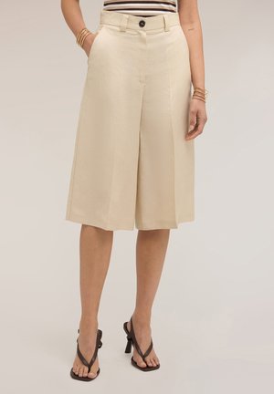 Frau trägt beige weite Shorts bis zum Knie, kombiniert mit schwarzen Sandalen mit Absatz und goldenen Armbändern, steht vor neutralem Hintergrund.