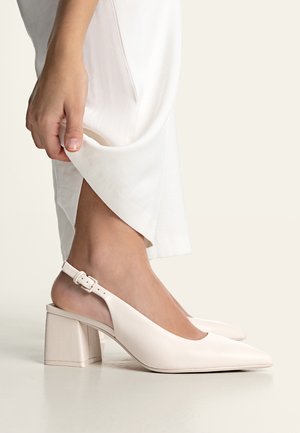 Vrouw die een witte enkellaars met blokhak met enkelbandje verstelt, terwijl ze witte broek draagt, staand op een gladde lichtgekleurde vloer.
