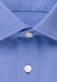 Gros plan sur le col d'une chemise bleue et deux boutons blancs, montrant la texture détaillée du tissu et les coutures.
