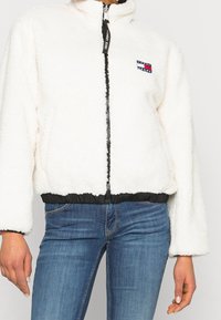 Biała, puszysta polarowa kurtka z czarnym zamkiem i kołnierzem, z delikatnym elastycznym wykończeniem na dole oraz małą łatą z logo na lewej piersi.