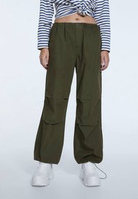 Pantalons large olive en vert avec une taille à cordon, dotés de grandes poches cargo et d'un tissu lisse et léger. Associés à des baskets blanches.