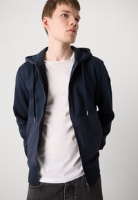 Navy zip-up hoodie with a drawstring hood, soft fabric, and contrasting lower panel; worn over a white shirt and gray jeans.