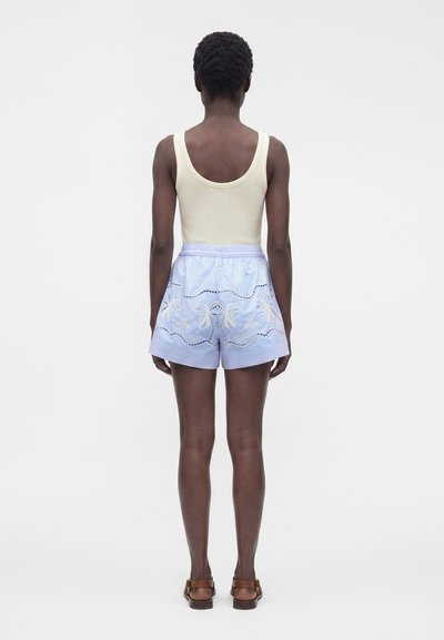 Personne aux cheveux courts et bouclés portant un haut beige sans manches, un short bleu clair brodé de palmiers et des sandales marron, debout de dos.