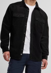 Chemise en velours côtelé noir à manches longues, boutonnée sur le devant, avec deux poches poitrine et une texture fine côtelée. Portée par-dessus un t-shirt blanc.