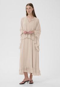 Beige dlouhé šaty s volány na rukávech a lemu, vyrobené z lehkého materiálu, s výstřihem do V a splývavým designem. Nosené černé sandály na tenkých páscích.
