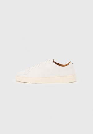 White low-top sneaker with lace-up design, minimal stitching details, and a cream-colored sole on a light gray background.