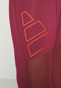 Bordeaux sportleggings met mesh panelen, voorzien van oranje geometrische accenten aan de zijkant en contrasterende stiksels.