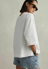 Hvid bomulds t-shirt med 3/4 ærmer, afslappet pasform, sideslids, parret med blå denim shorts. Modellen bærer mørke solbriller.