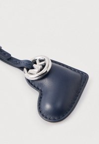 Tamnoplavi kožni privjesak za ključeve u obliku srca sa srebrnim metalnim dijelovima. Ima sjajnu teksturu i šavove duž rubova.