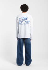 Biała koszulka z długim rękawem z niebieską grafiką przedstawiającą tacos i napoje na plecach. Luźne niebieskie dżinsy z szerokimi nogawkami.