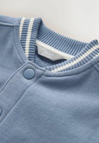 Jasnoniebieska bluza z prążkowanym kołnierzem ozdobionym białymi paskami, zapinana na guziki, z małą metką z napisem „NEXT”. Miękka w dotyku.