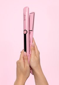 Roze haarstijlijzer vastgehouden met twee handen, met een glanzende afwerking, zwarte accenten, rechthoekige platen en een ronde aan/uit-knop.