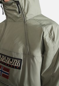 Olivgrön nylonpulloverjacka med hög krage, framzipper, svart logotypapplikation och en norsk flagga som accent. Slät textur genomgående.