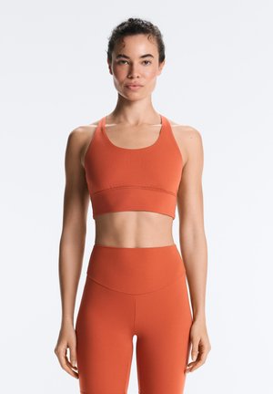 Edzett nő áll szemből, felül rozsdabarna sportmelltartót és magas derekú leggingset visel, egyszerű háttér előtt.