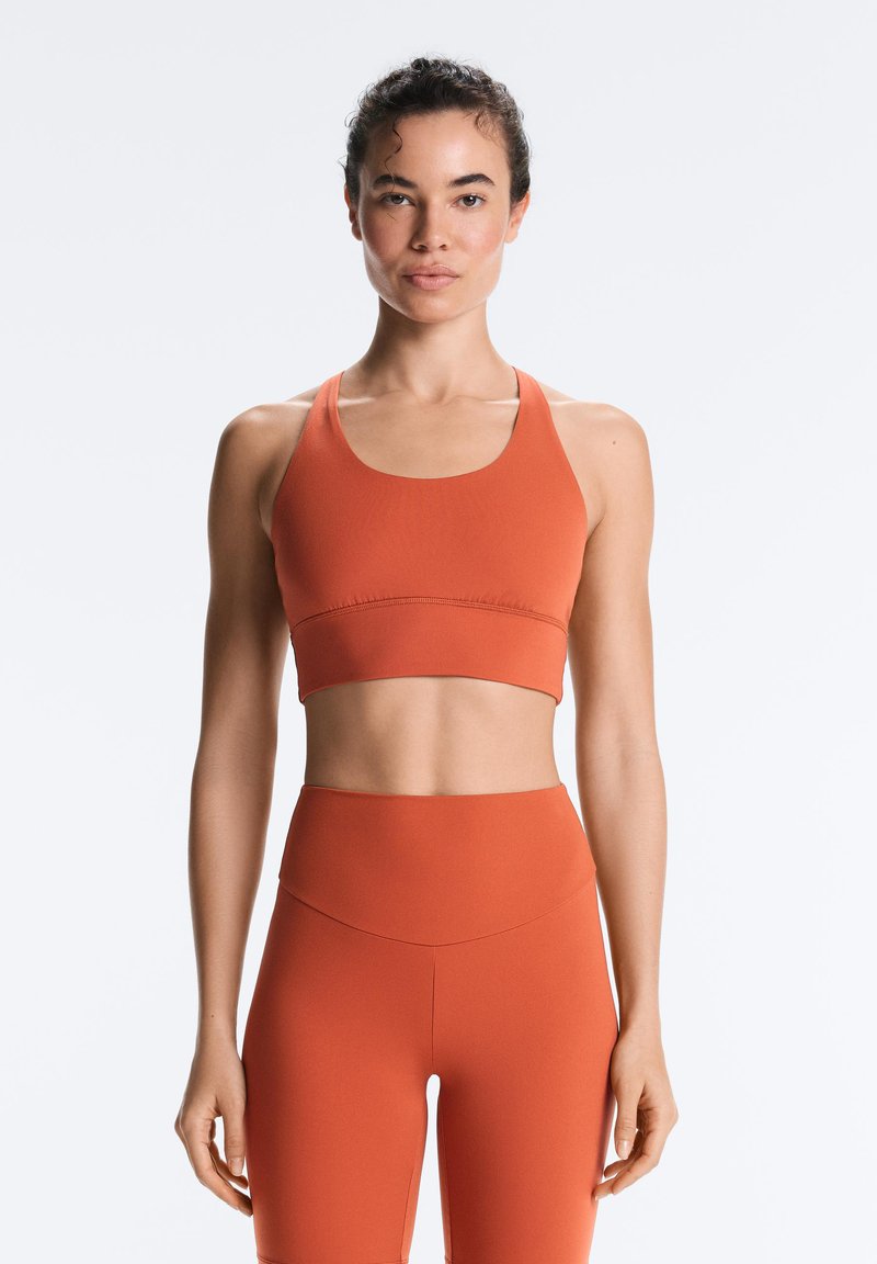 Fit woman stands facing forward wearing matching rust-orange sports bra and high-waisted leggings against a plain background.