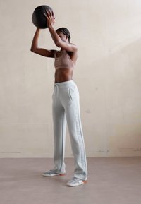 Sporta apģērbs, kas sastāv no beigu krāsas sporta krūštura un gaiši zilu sporta biksēm ar sānu tekstu. Cilvēks tur melnu medicīnas bumbu.