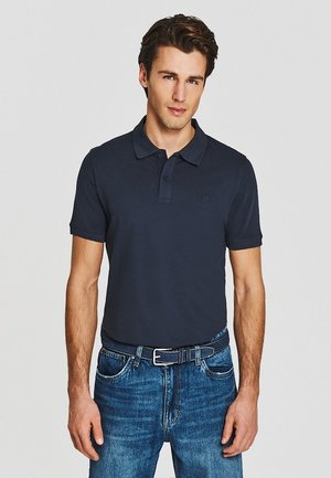 Młody mężczyzna ubrany w ciemnogranatową koszulkę polo wpuszczoną w niebieskie dżinsy z czarnym paskiem, stojący na jednolitym jasno szarym tle.