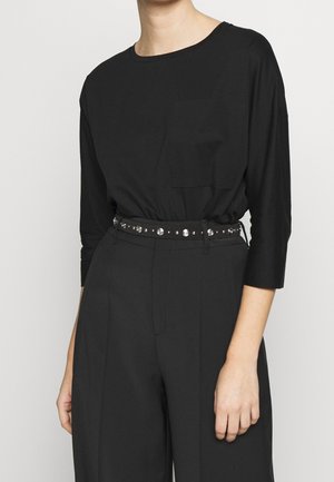 Personne portant une chemise noire à manches longues avec une poche poitrine rentrée dans un pantalon noir taille haute avec une ceinture noire cloutée.