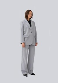 Completo grigio su misura con giacca a un bottone e pantaloni a gamba larga. Tessuto liscio con tasche, indossato sopra una camicia nera. Scarpe nere a punta.