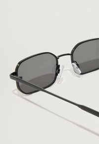 Čierne obdĺžnikové slnečné okuliare so šedým tónovaným sklom, tenkým kovovým rámom a priehľadnými nosovými podložkami. Dizajn obsahuje jemné drážky ako ozdobu.