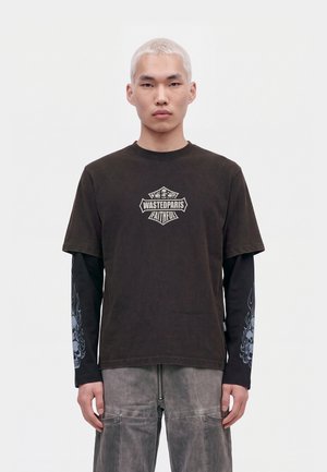 Un mannequin aux cheveux courts décolorés porte un T-shirt foncé à motif par-dessus un haut imprimé à manches longues et un pantalon cargo gris, sur fond blanc.