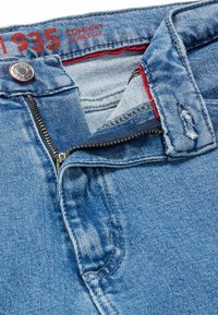 Jeansen i ljusblå denim har en dragkedja. Funktionerna inkluderar en silverknapp, röd sömnad och ett bekvämt stretchtyg.