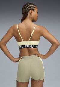 Sostén deportivo amarillo claro con banda negra, con el logo de PUMA. Pantalones cortos verde oliva con acentos laterales, textura suave, diseño ajustado.