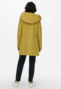 Senfgelber Wollmischmantel mit Kapuze, mittellangem Schnitt, strukturierter Oberfläche und gerader Silhouette. Getragen mit dunkler Hose und weißen Sneakers.