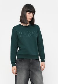 Donna con capelli scuri alla spalla che indossa un maglione verde scuro con "PARIS" ricamato e jeans neri sbiaditi, in piedi contro uno sfondo bianco.