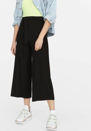 Femme portant un pantalon large noir raccourci avec une ceinture à la taille, un haut vert clair, une veste en denim clair et des baskets blanches sur un fond blanc.