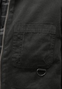 Czarna kurtka z materiału typu canvas, wyposażona w kieszeń na piersi z klapką, metalowy kółko D oraz zamek błyskawiczny z widocznymi detalami szwów.