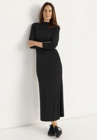 Musta pitkähihaine mekko, jossa on mock neck, tiukka siluetti ja sivu rypytys. Yhdistetty mustiin lenkkareihin, joissa on valkoisia yksityiskohtia.