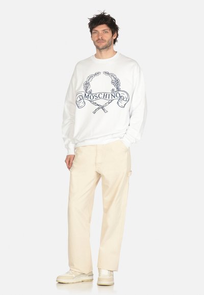 Felpa bianca con logo "Moschino" blu, una corona decorativa e elementi incrociati. Abbinata a pantaloni beige oversize e scarpe di colore chiaro.