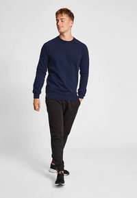 Marineblauer Pullover mit langen Ärmeln und Rundhalsausschnitt, kombiniert mit schwarzen Jogginghosen und Sportschuhen, mit lockerem Schnitt.