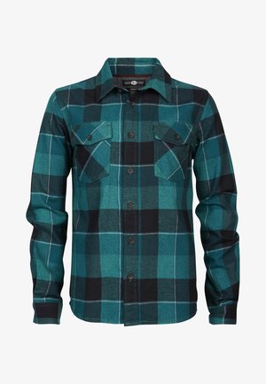 Osoba ubrana w zieloną i czarną koszulę w kratę, niebieskie dżinsy i czarne sportowe buty za kostkę, stoi z nogami szeroko rozstawionymi.