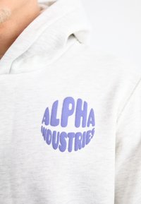 Šviesiai pilkas džemperis su pūkuotu violetiniu apvaliu logotipu, ant kurio yra užrašas "ALPHA INDUSTRIES", išsiskiriantis minkšta medžiaga ir laisvu siluetu.