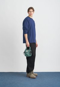 Laivastonsininen pitkähihaisessa paidassa, mustissa housuissa, harmaissa saappaissa, kantaen vihreää laukkua, jossa on logo. Yksinkertainen muotoilu, rento istuvuus ja neutraalit värit.