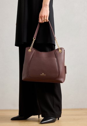 Femme en pantalon large noir et bottes tenant un sac à main en cuir marron foncé avec des anses chaîne dorées de la marque Anna Field.