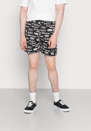 Homme portant un short imprimé logo noir et blanc, des chaussettes blanches, des baskets noires et un t-shirt blanc, debout devant un fond uni.