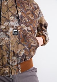 Camo-gepatroniseerde zip-up jas in bruin, beige en groene tinten; voorzien van een zwart logopatch en een bruine ribgebreide zoom; gestructureerde stof.
