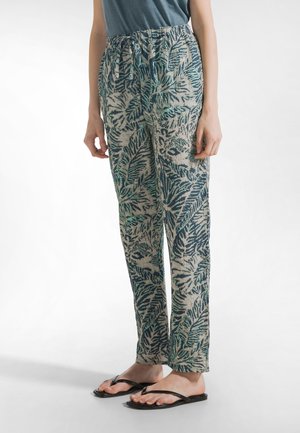 Linen blend trousers with a leafy green and beige pattern, featuring an elastic waistband and relaxed fit. Paired with black flip-flops.