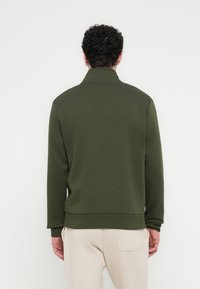 Σκούρο πράσινο φούτερ με ψηλό γιακά, ριμπ μανσέτες και εφαρμοστό τελείωμα. Το υλικό φαίνεται μαλακό με ομαλή υφή.