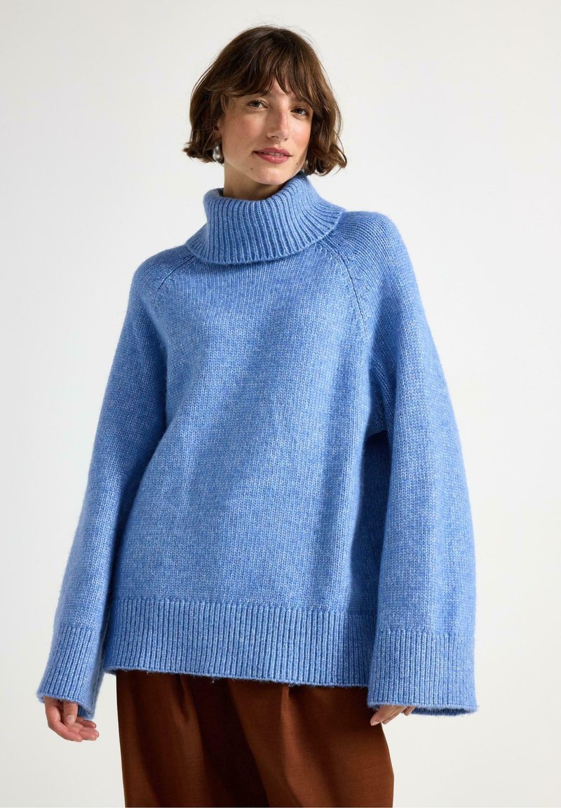 Kvinde iført en oversized lyseblå turtleneck sweater og løse brune bukser foran en ensfarvet hvid baggrund.