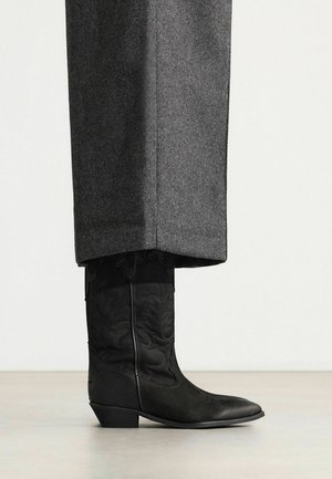 Zwarte leren cowboylaars met decoratief stiksel, gedragen onder lange grijze wollen broeken tegen een egale lichte achtergrond.
